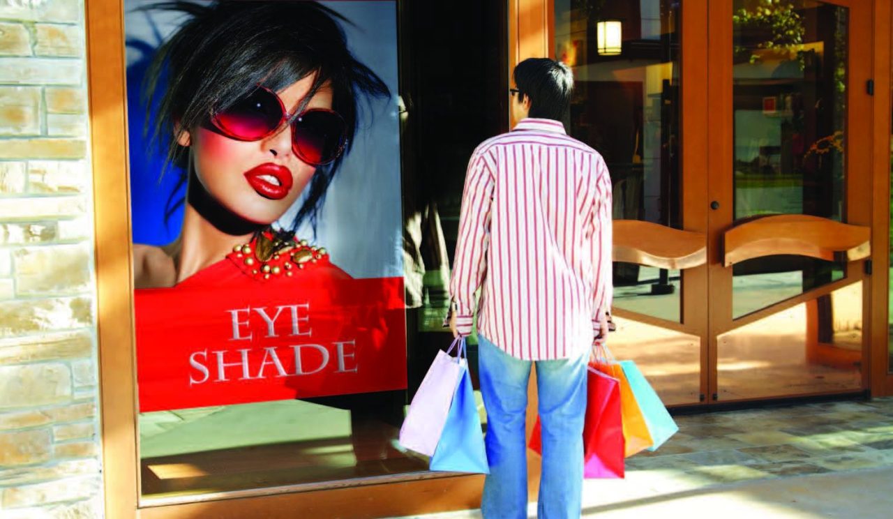 Man standing in front of a store looking at a window graphic of a model wearing Eye Shade sunglasses.