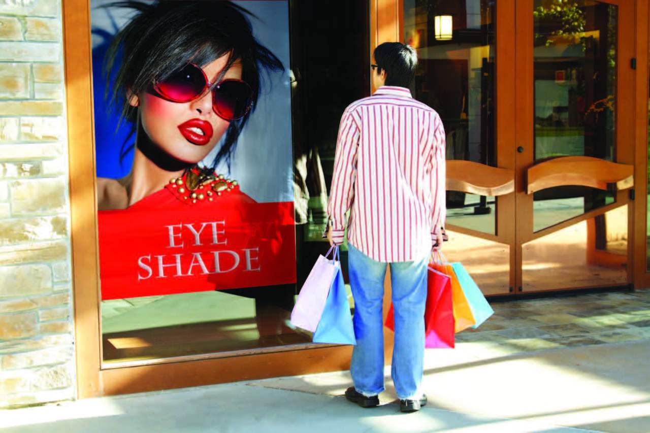 Man standing in front of a store looking at a window graphic of a model wearing Eye Shade sunglasses.